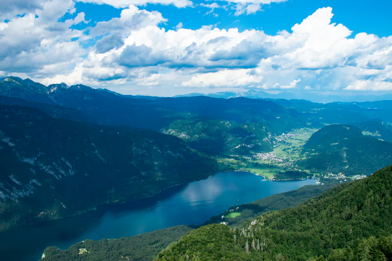 Vue en hauteur sur un lac bleu bordé de forêts, devant plusieurs chaînes de montagnes sous un ciel nuageux. Photographie d’illustration.